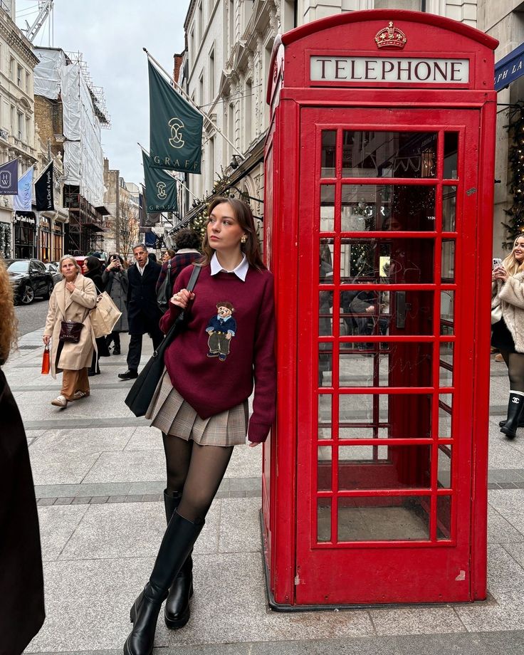 Woman in a plaid skirt and long boots leaning against a red telephone booth on a city street