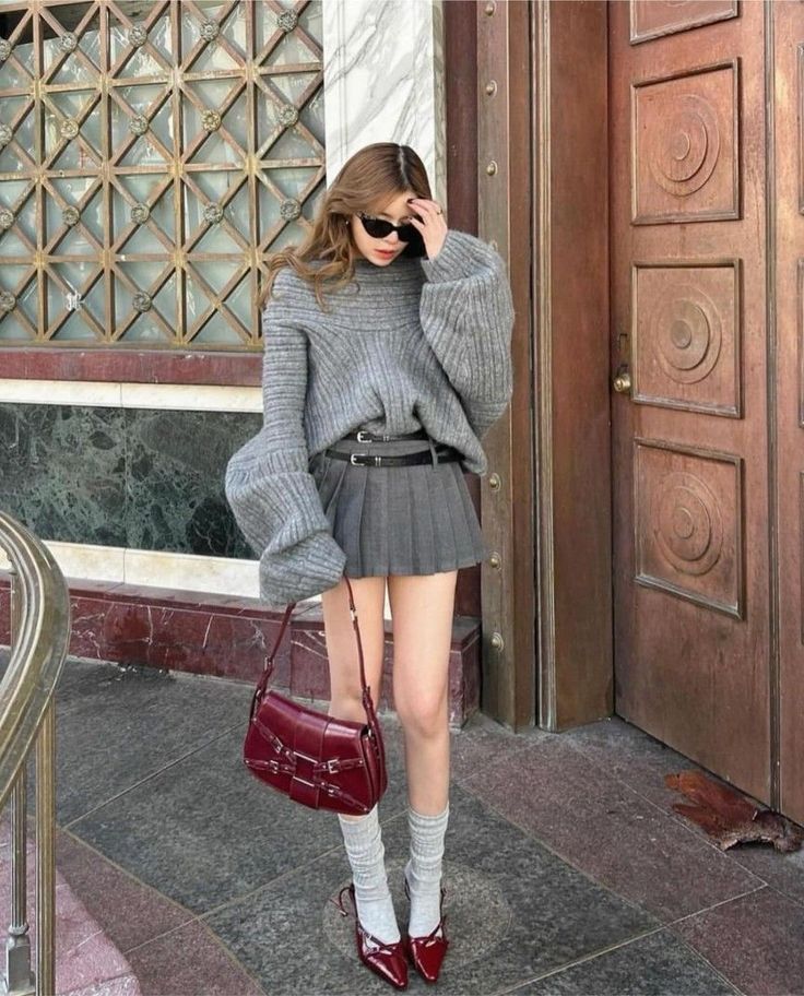 Woman in a gray sweater and pleated skirt with a red handbag and heels