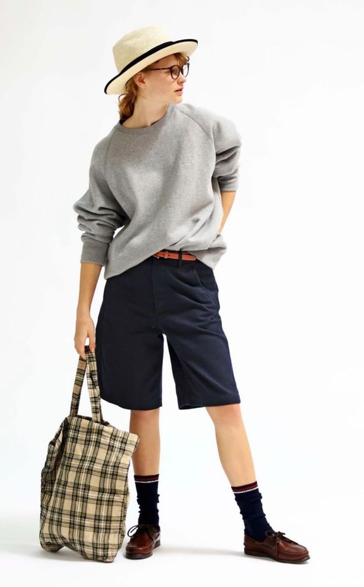 Person in a straw hat, glasses, grey sweatshirt, and navy shorts holding a plaid tote bag