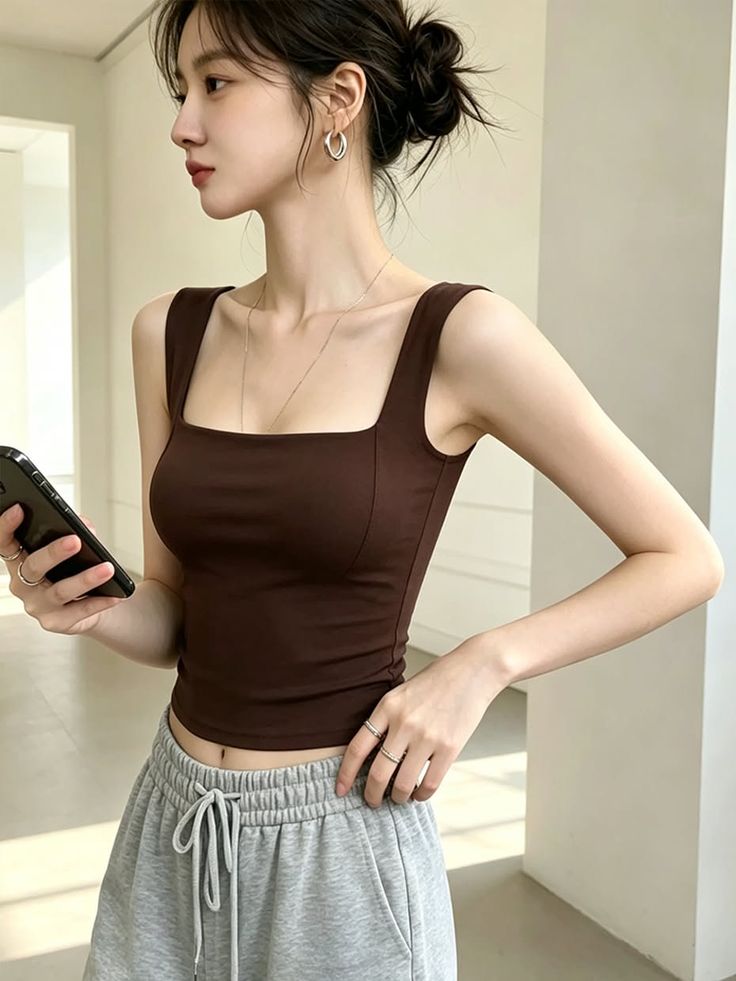 Woman in a brown tank top and grey sweatpants holding a phone indoors