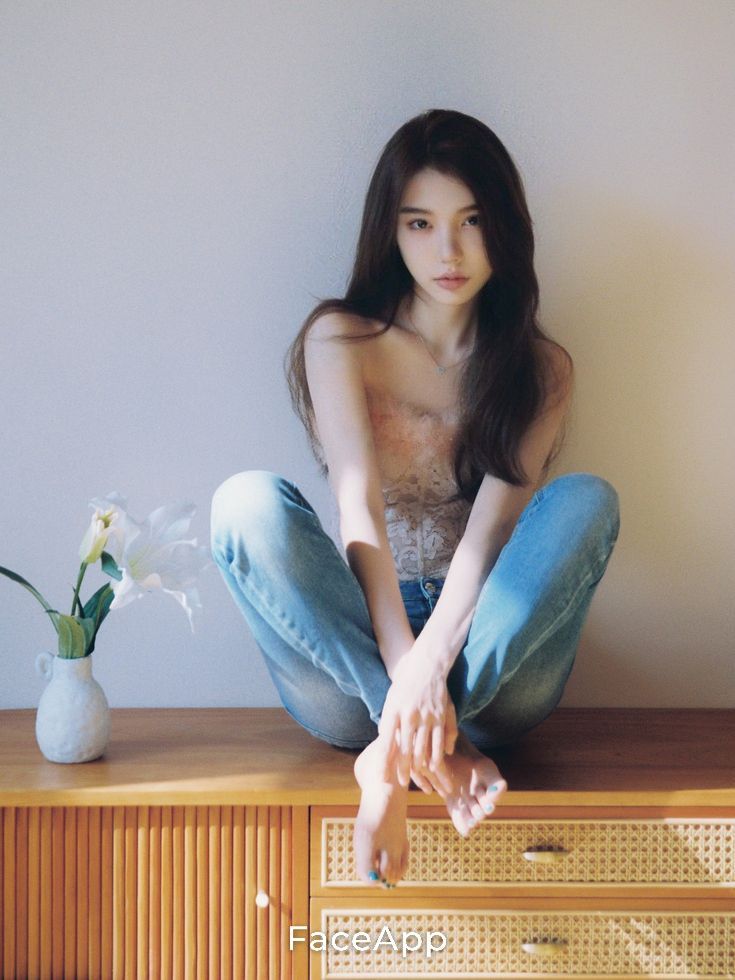 Woman with long hair wearing jeans and a strapless top, sitting on a wooden cabinet