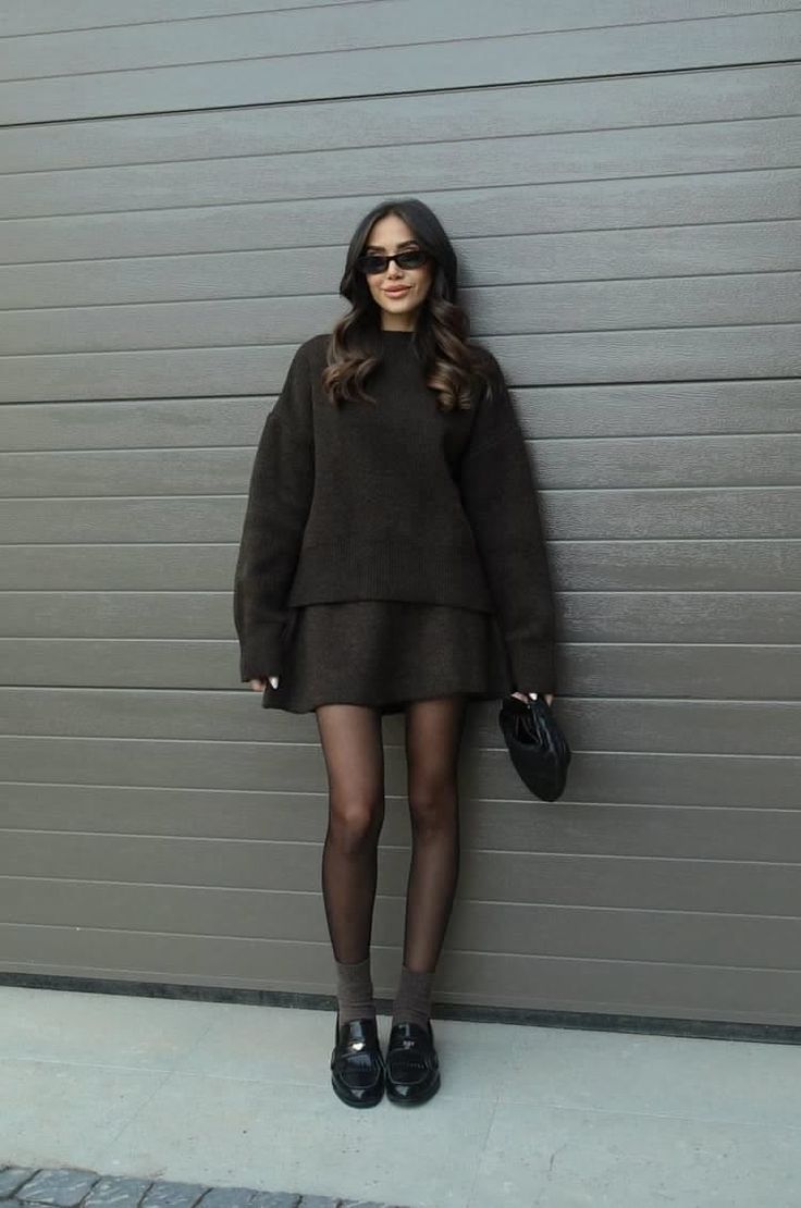 Woman in a dark sweater and mini skirt with sunglasses leaning against a wall