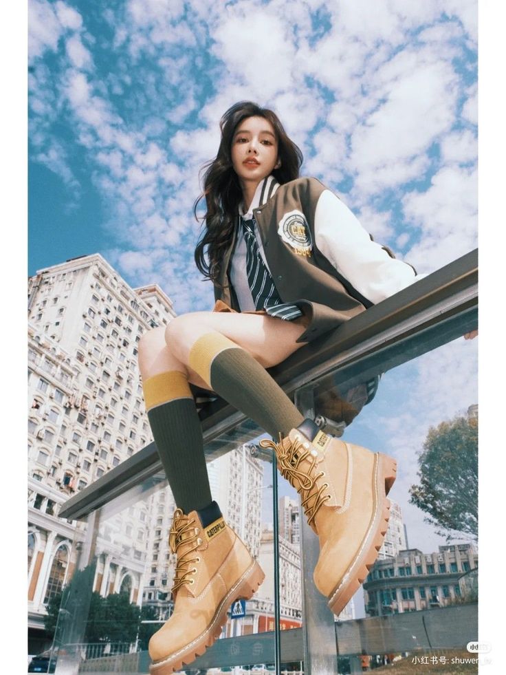 Woman in a varsity jacket and combat boots sitting on a ledge against a blue sky
