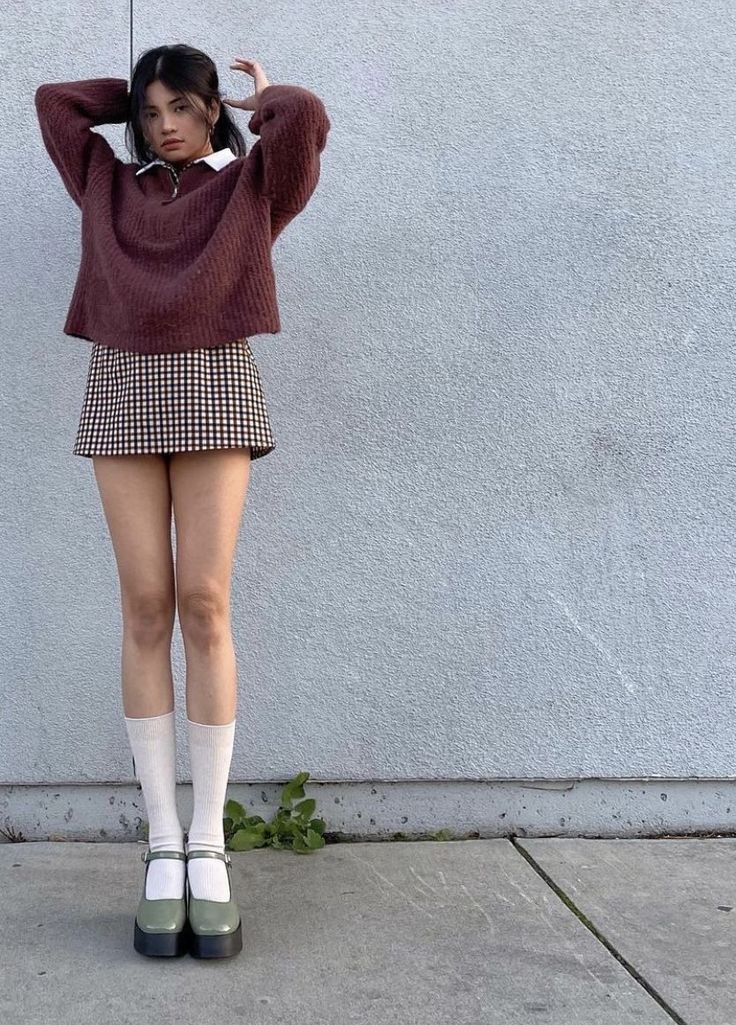 Woman in a brown sweater and gingham skirt posing against a wall