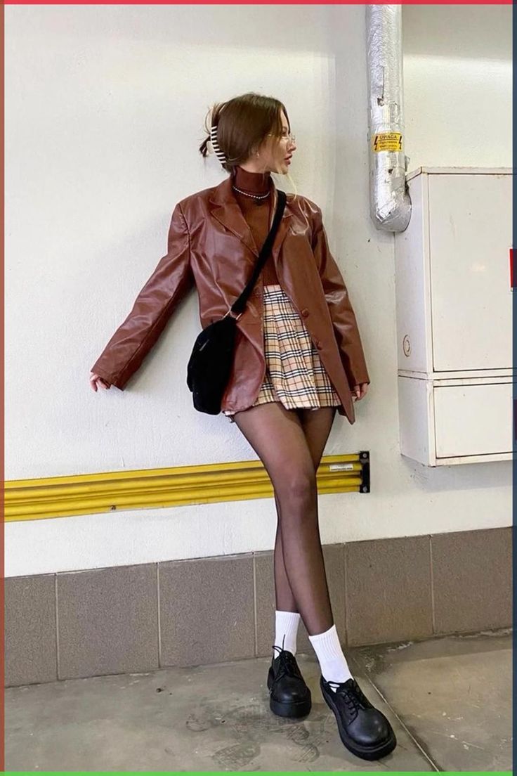Woman in a brown leather jacket and plaid skirt leaning against a wall