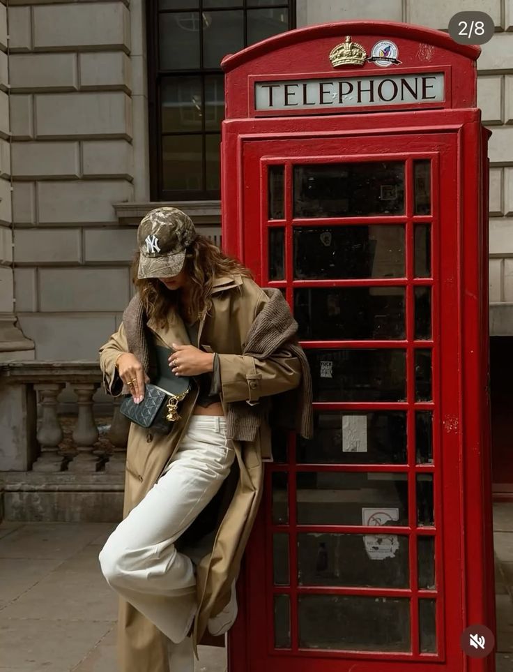 Woman in a trench coat and baseball cap leaning on a red telephone booth