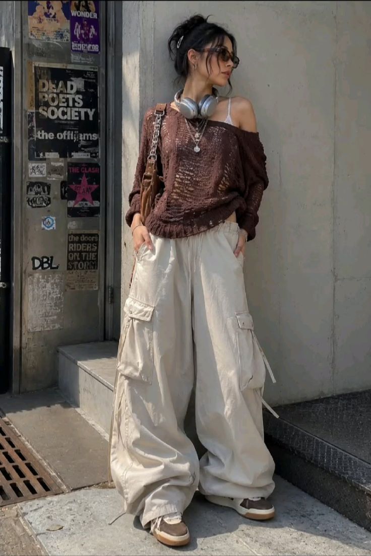 Woman in a loose knit sweater and baggy cargo pants leaning against a wall outdoors