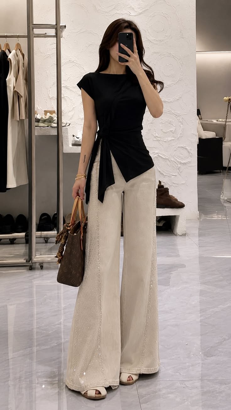 Woman in a black top and wide leg pants taking a mirror selfie indoors
