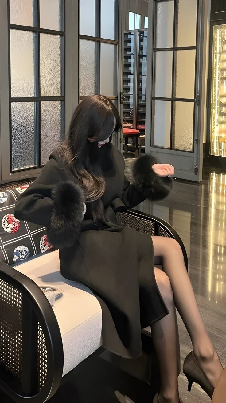 Woman in a black coat with fur cuffs and black tights sitting indoors