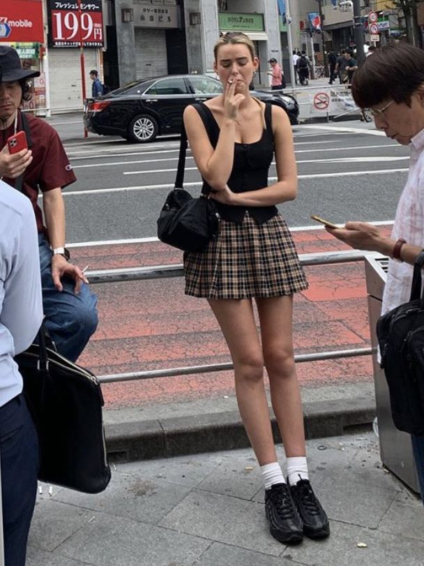 Woman in a plaid skirt and black top smoking on a city street