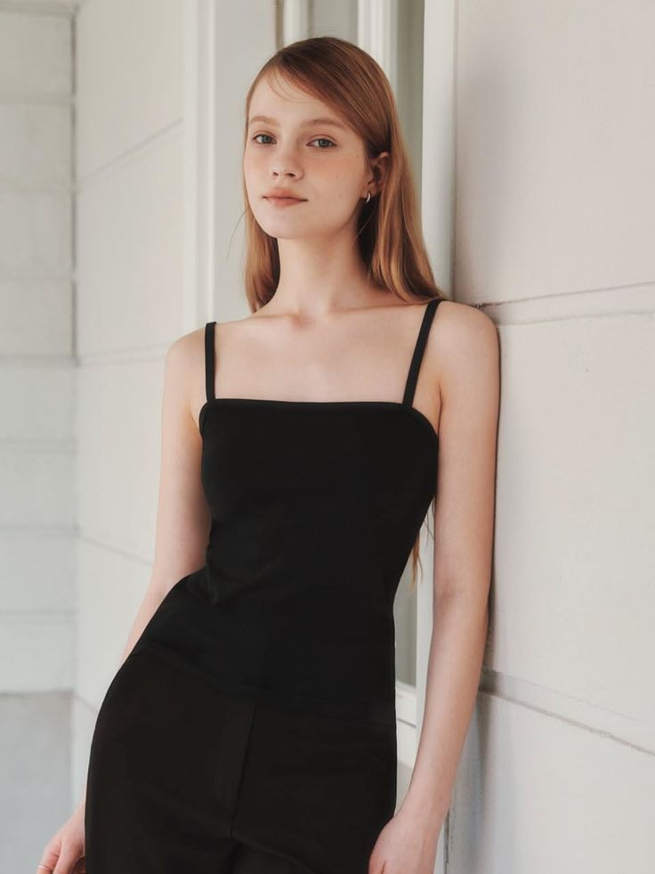 Young woman with long hair wearing a black top leaning against a white wall