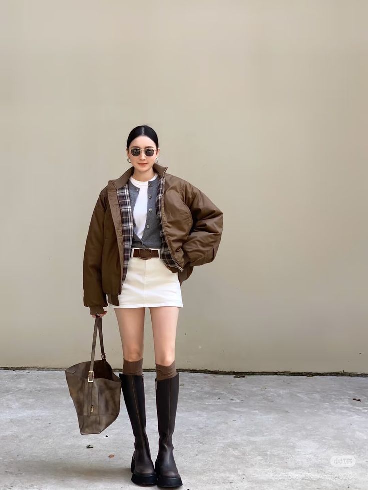 Woman in a brown jacket, white mini skirt, and knee high boots standing outdoors