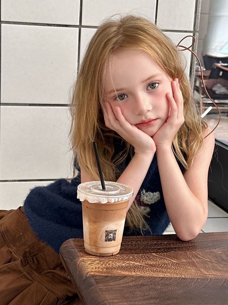 Young girl with long blonde hair resting her chin on her hands next to an iced coffee