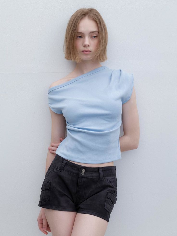 Woman with short blonde hair wearing a blue off-shoulder top and black shorts