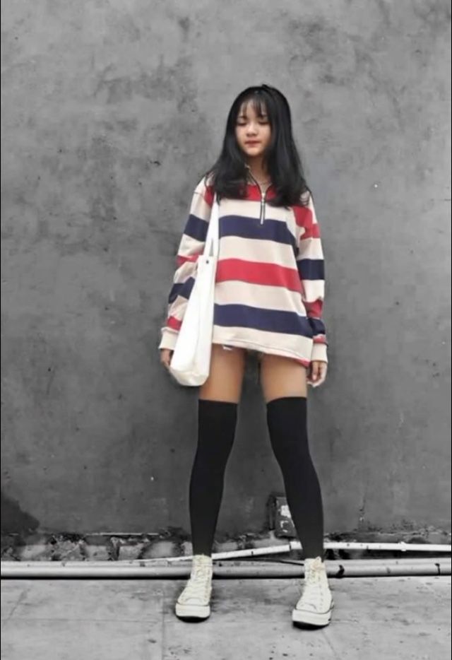 Young woman in a striped shirt, thigh-high socks, and sneakers against a gray wall