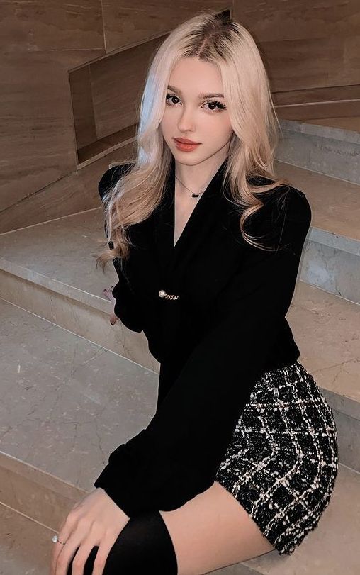 Woman with blonde hair wearing a black blazer and tweed skirt indoors