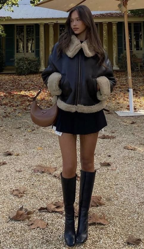 Woman in a shearling jacket and black skirt standing outdoors with autumn leaves