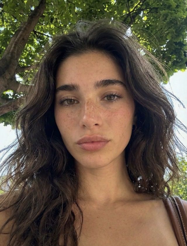 Woman with long brown hair and freckles outdoors under green trees