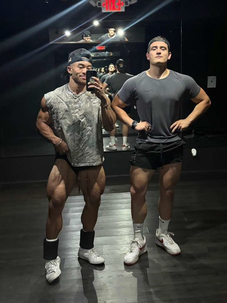 Two muscular men posing in a gym mirror, one taking a selfie