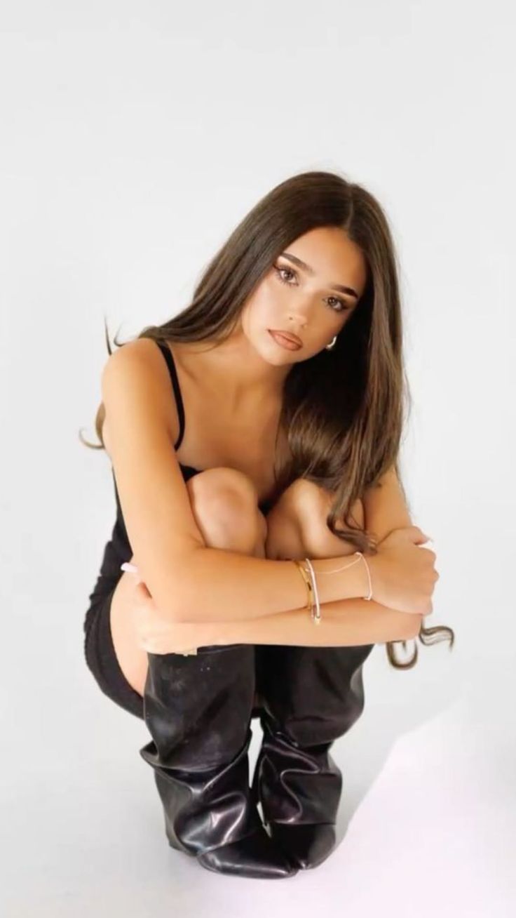 Woman with long brown hair in a black dress and boots squatting in a studio