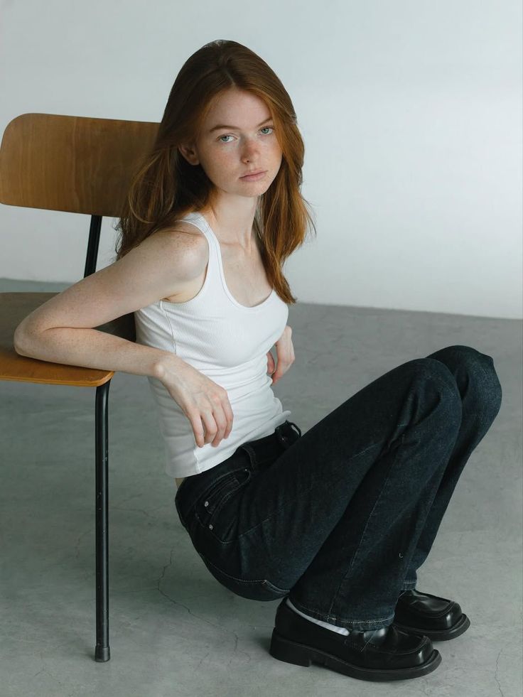 Woman with red hair and freckles wearing a white tank top and jeans, sitting on the floor