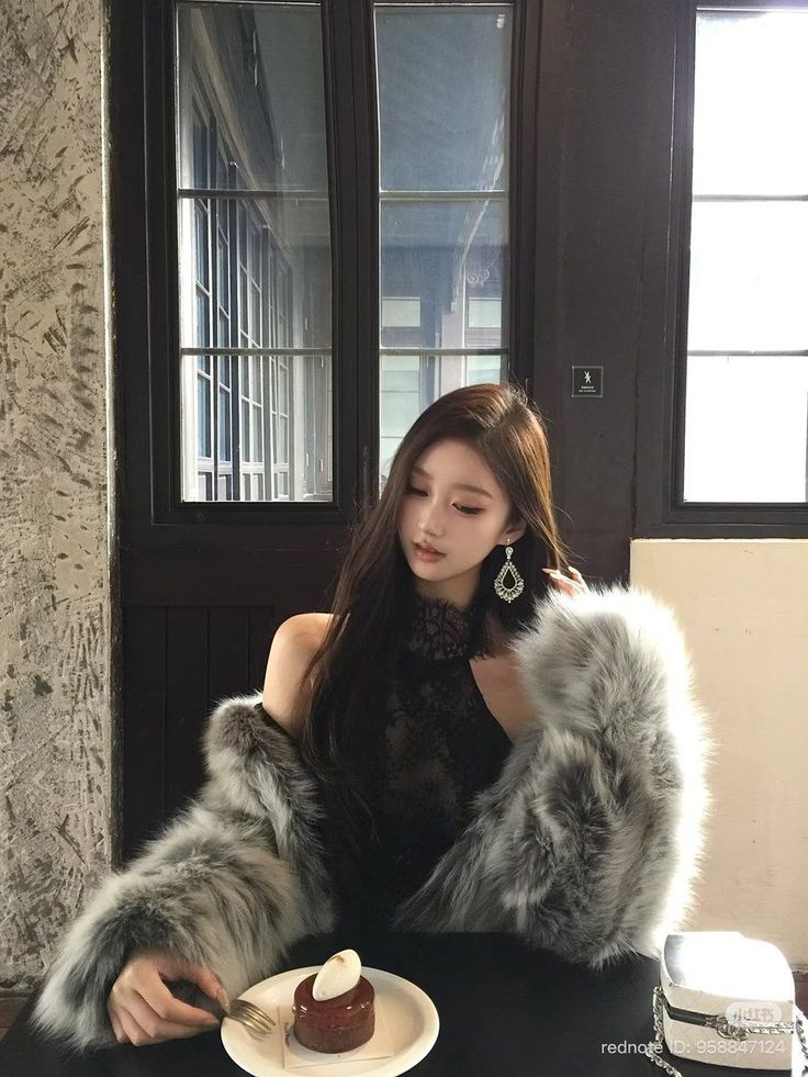 Woman with long hair wearing a fur coat and black lace top, eating dessert indoors