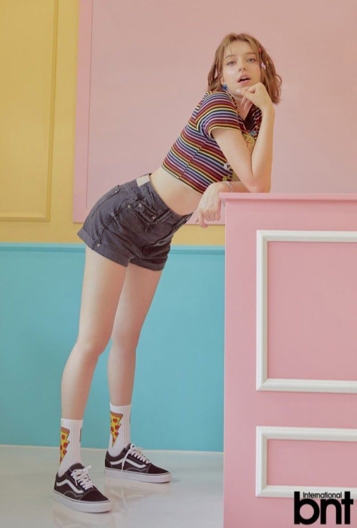 Woman in a rainbow crop top and denim shorts leaning on a pink surface