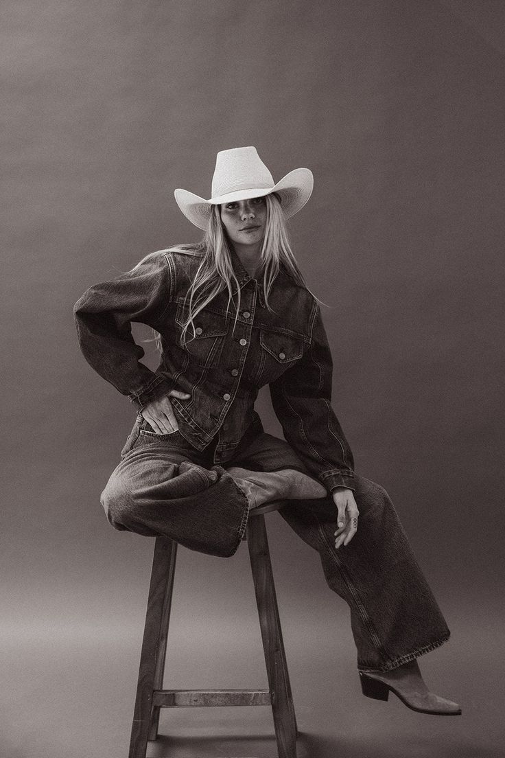 Woman in a cowboy hat and denim outfit sitting on a stool in a studio