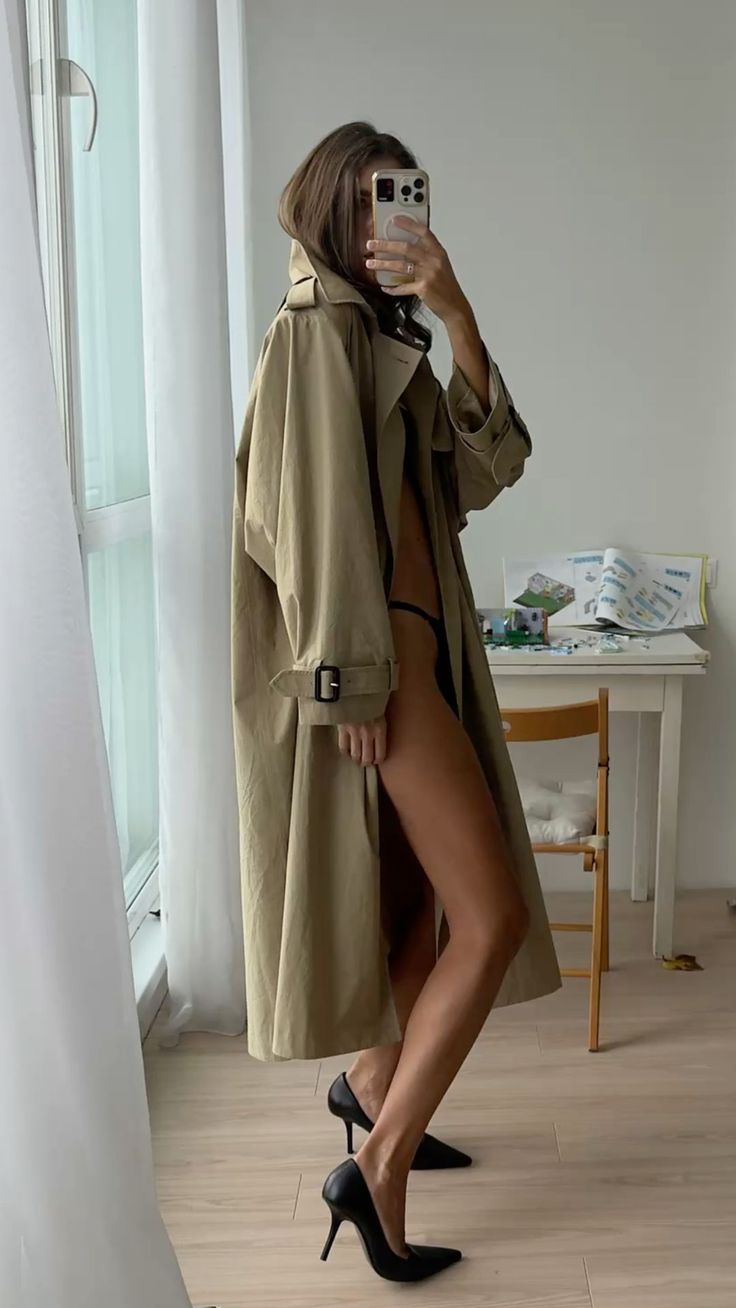 Woman in a trench coat and black heels taking a mirror selfie