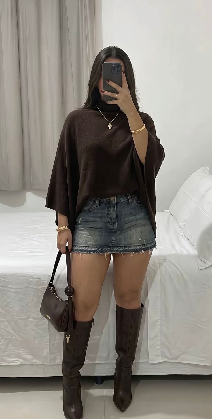 Woman in a brown top, denim skirt, and brown boots holding a handbag indoors
