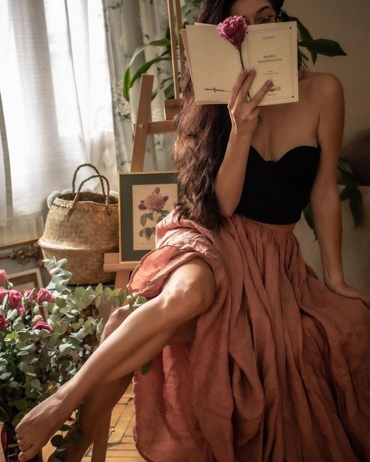 Woman with long hair holding a book and a flower, wearing a strapless top and long skirt