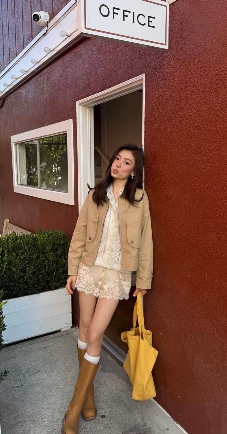 Woman in a tan jacket and lace shorts leaning against a red wall with a yellow tote bag
