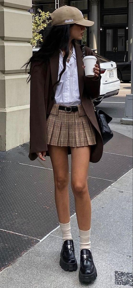 Woman in a plaid skirt, brown blazer, and baseball cap on a city street