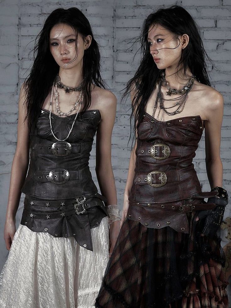 Two women in steampunk-inspired leather corsets and long skirts against a white brick wall