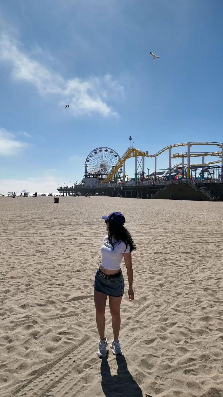 Woman in a denim skirt and crop top on a beach with an amusement park in the background