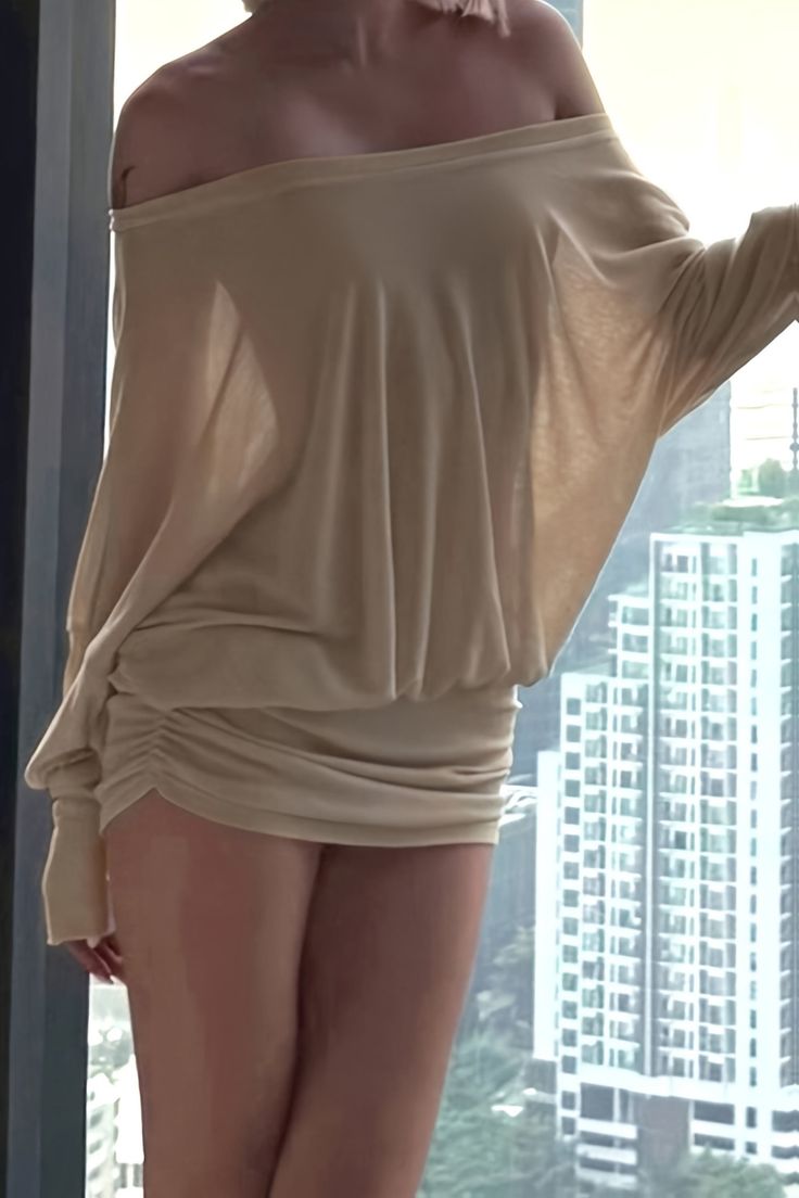 Woman in a beige off-shoulder dress standing by a window overlooking a cityscape
