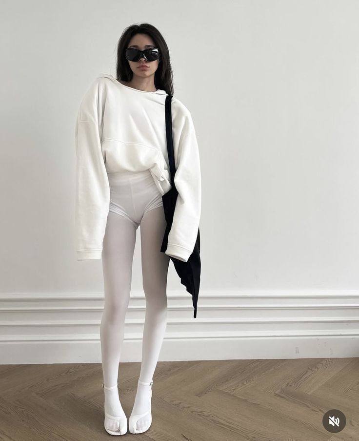 Woman in a white hoodie, white tights, and black sunglasses standing indoors