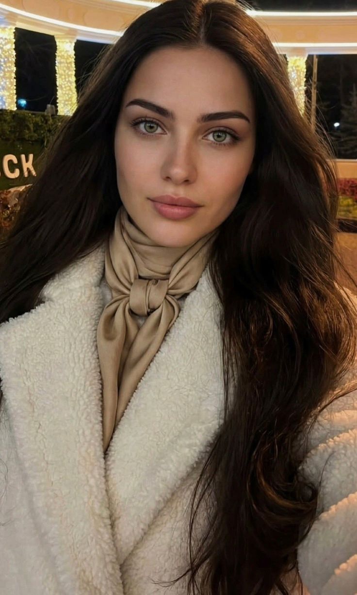 Woman with long dark hair and green eyes wearing a white coat and beige scarf at night