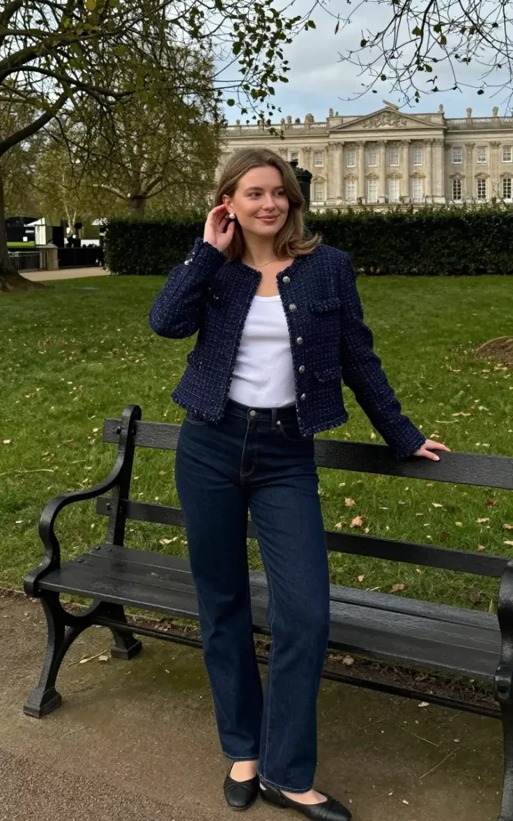 Woman in a tweed jacket and jeans standing by a park bench with a building in the background