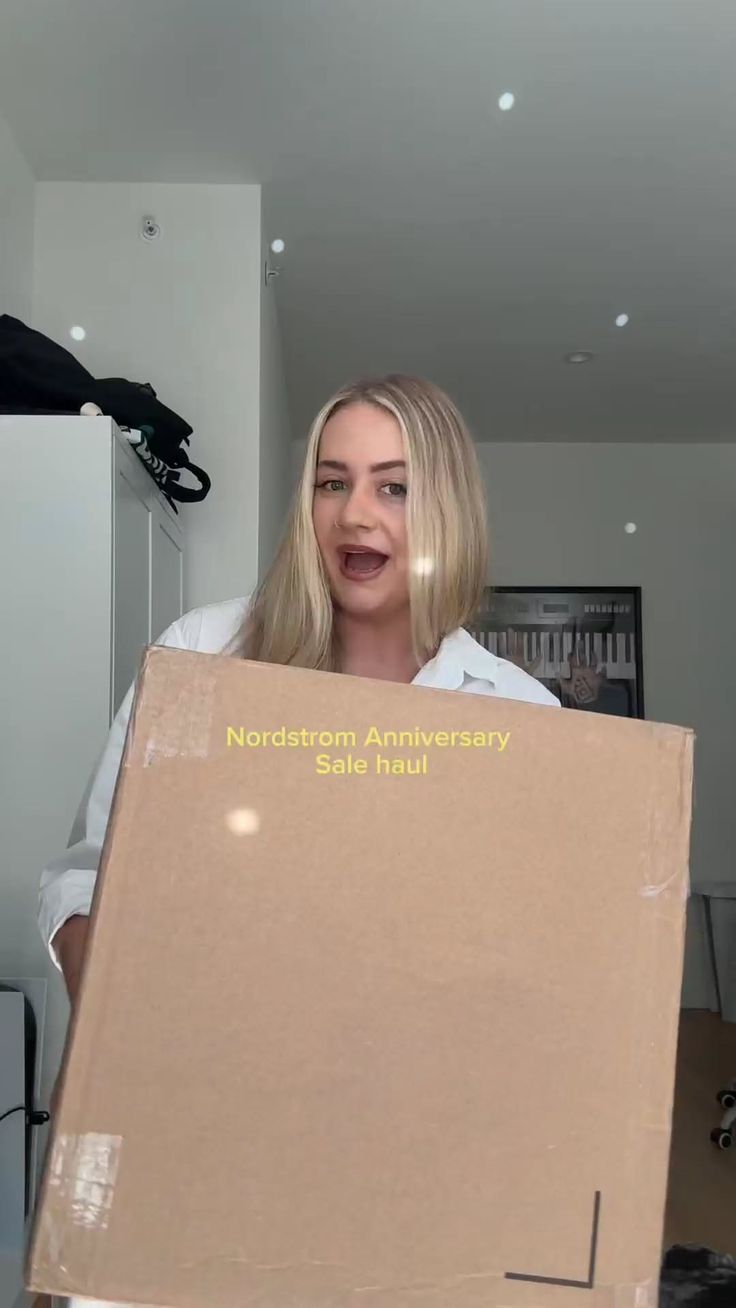 Woman with blonde hair holding a Nordstrom Anniversary Sale haul box indoors