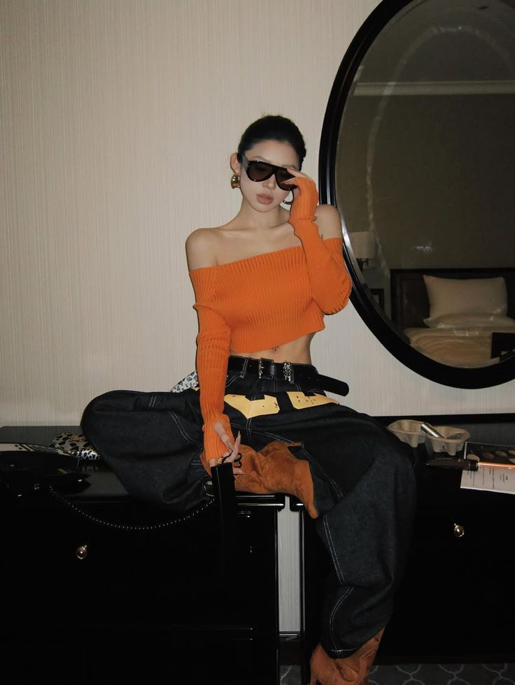 Woman in an orange off-shoulder top and baggy pants sitting on a dresser