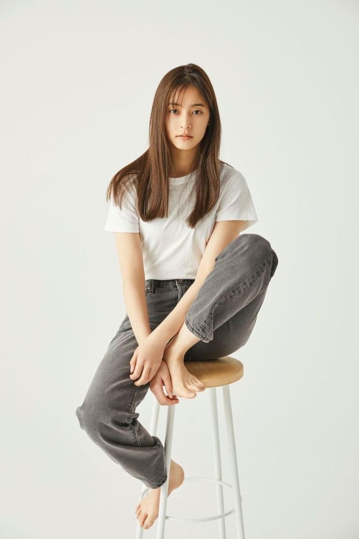 Woman with long hair in a white t-shirt and gray jeans sitting barefoot on a stool