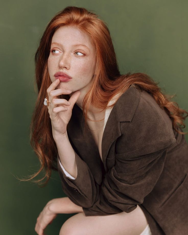 Woman with red hair and freckles wearing a brown jacket against a green background