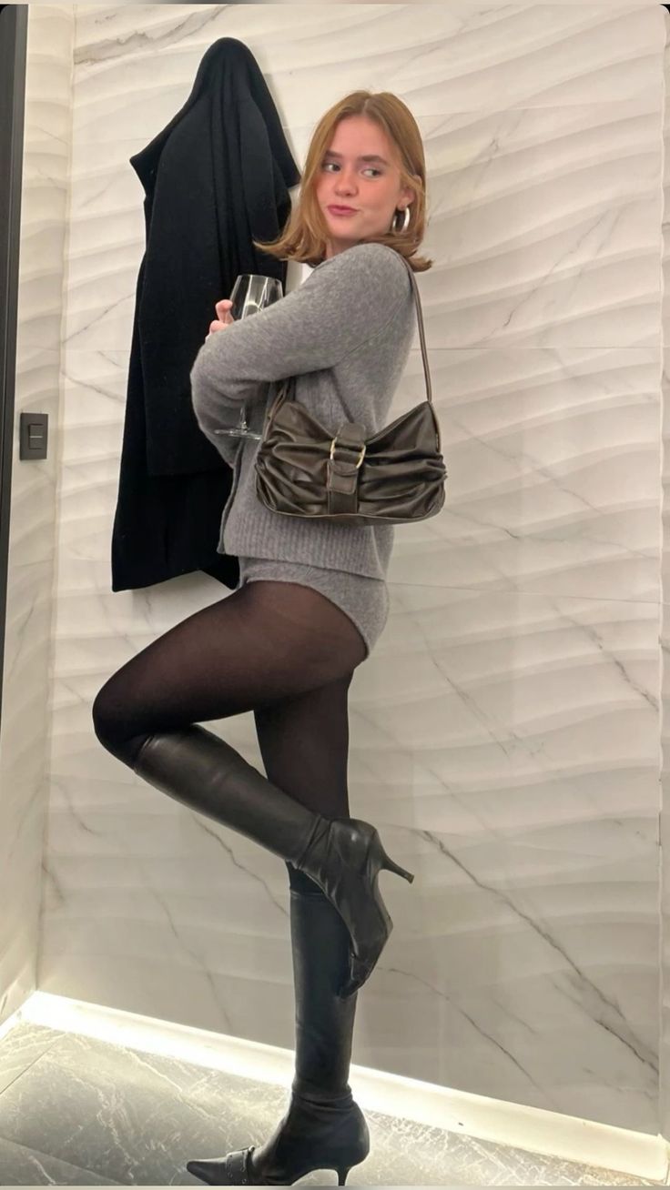 Woman in a grey cardigan and black tights posing indoors with a glass of wine