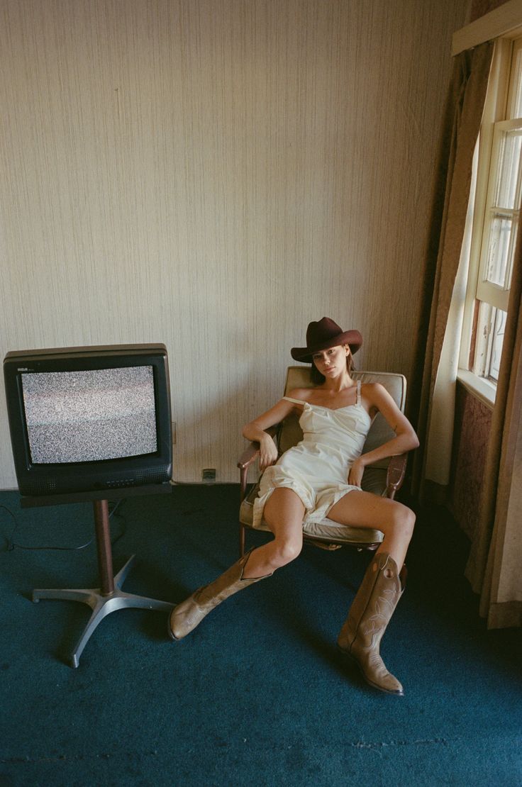 Woman in a cowboy hat and slip dress sitting in a chair next to a static TV
