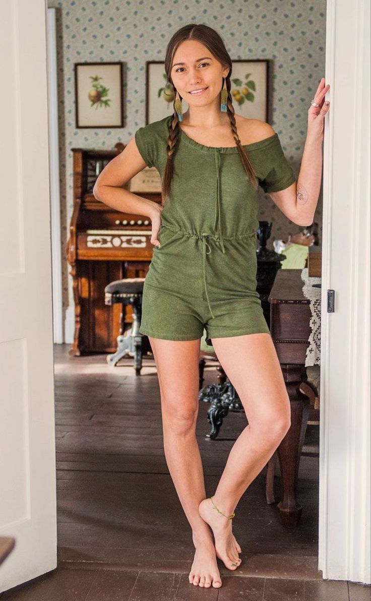 Woman in a green romper with braided hair standing barefoot in a doorway