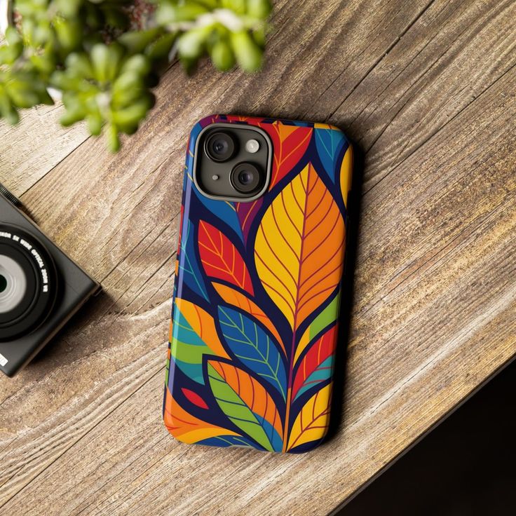 A phone case with a colorful leaf pattern on a wooden table next to a camera
