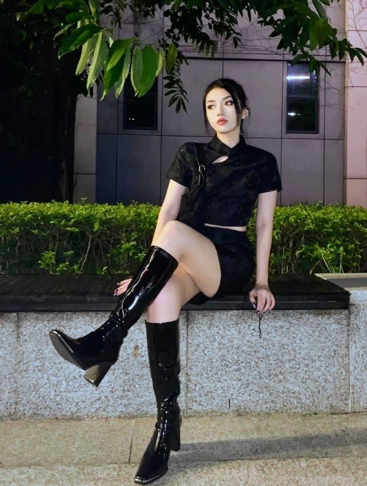 Woman in a black outfit and knee high boots sitting outdoors at night