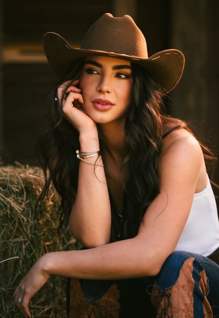Woman in a cowboy hat and white tank top posing outdoors with hay bales