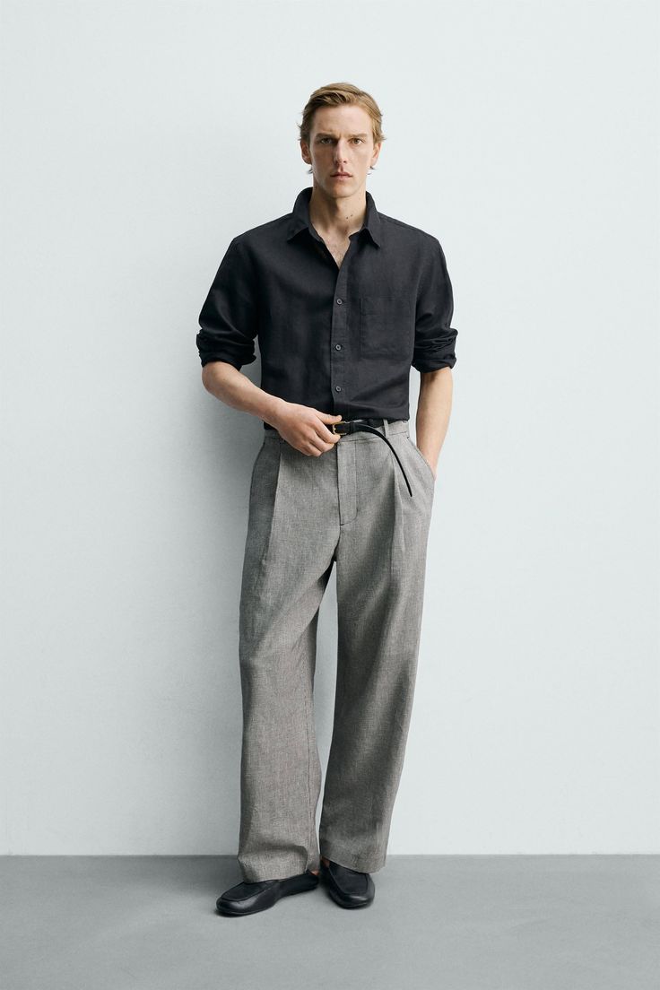 Man in a black shirt and grey pants posing against a white wall